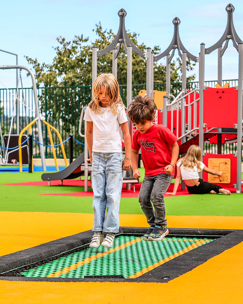 Children jump on a colorful inground trampoline at a playground.